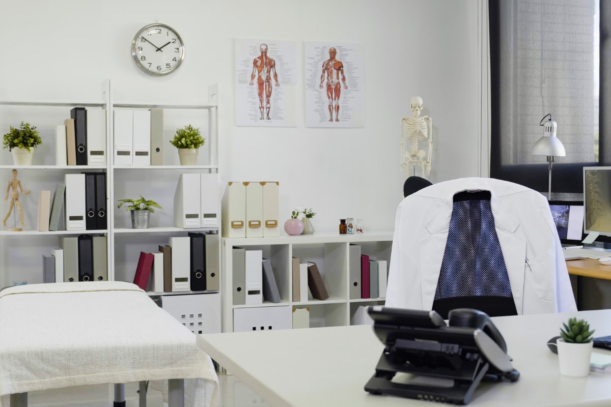 interior of a doctors office with a desk, white coat hanging from a chair and a treatment table