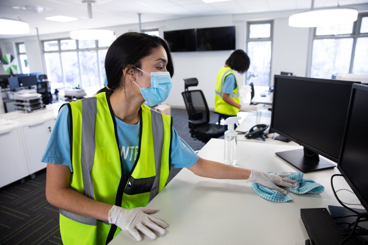 woman in face mask cleaning office desk top with another cleaning woman in the background